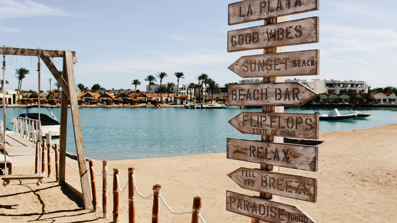Holzwegweiser an einem Sandstrand in Hurghada, dahinter Boote und Meer